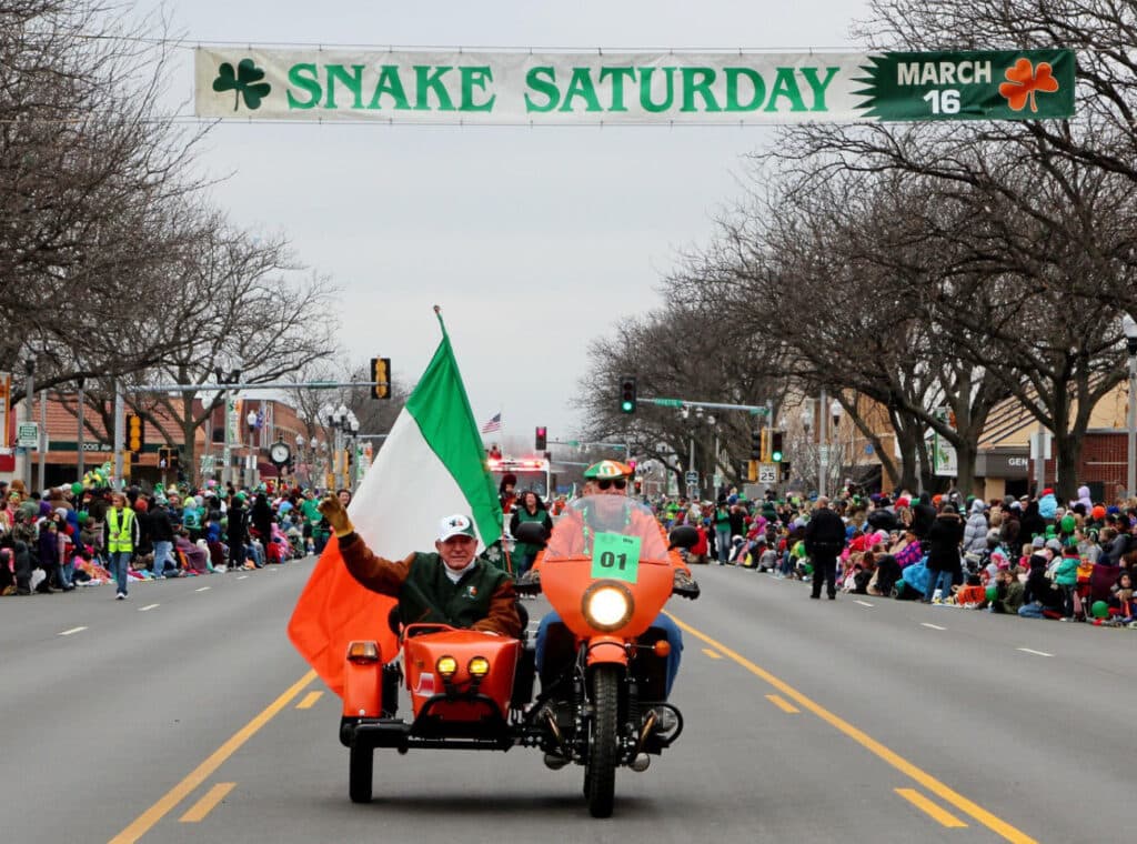 Mickey Finn waving from an orange motorcycle sidecar with an Irish flag, leading the parade under a Snake Saturday banner